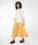 model in a white top with a hankerchief hem and hip pocket, worn with a buttery yellow pant with a drape detail on the sides